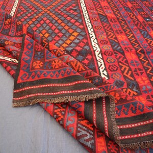 Pu&ograve; includere: Un tappeto piegato con un vivace motivo geometrico rosso, nero e blu. Il design include forme a diamante e motivi ripetuti lungo i bordi. Il tappeto ha una frangia marrone scuro.