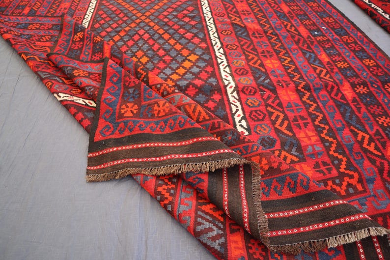 Pu&ograve; includere: Un tappeto piegato con un vivace motivo geometrico rosso, nero e blu. Il design include forme a diamante e motivi ripetuti lungo i bordi. Il tappeto ha una frangia marrone scuro.