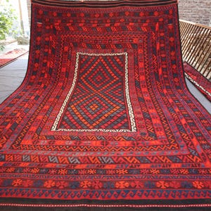 Pu&ograve; includere: Un grande tappeto rettangolare rosso con intricati motivi geometrici. Il tappeto presenta un motivo centrale a diamante nei toni del rosso, blu e arancione, incorniciato da un bordo bianco. Il bordo e i bordi sono adornati da dettagliati motivi tribali.