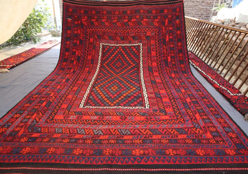 Pu&ograve; includere: Un grande tappeto rettangolare rosso con intricati motivi geometrici. Il tappeto presenta un motivo centrale a diamante nei toni del rosso, blu e arancione, incorniciato da un bordo bianco. Il bordo e i bordi sono adornati da dettagliati motivi tribali.