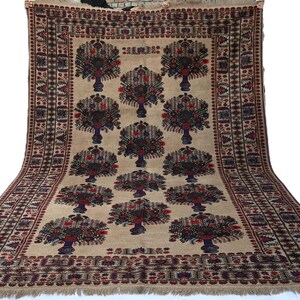 May include: Beige rug with a repeating pattern of stylized trees in vases. The trees have dark red and blue details, set against a beige background. The rug has a detailed border with geometric patterns and fringe.