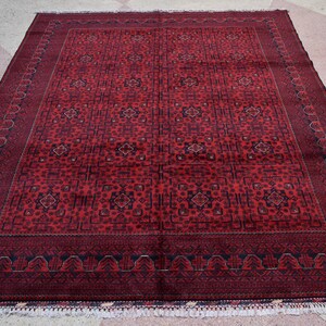 May include: A rectangular, hand-woven rug with a deep red field and intricate geometric patterns. The rug features a dark border and decorative fringe along the edges. The design includes repeating floral and geometric motifs.