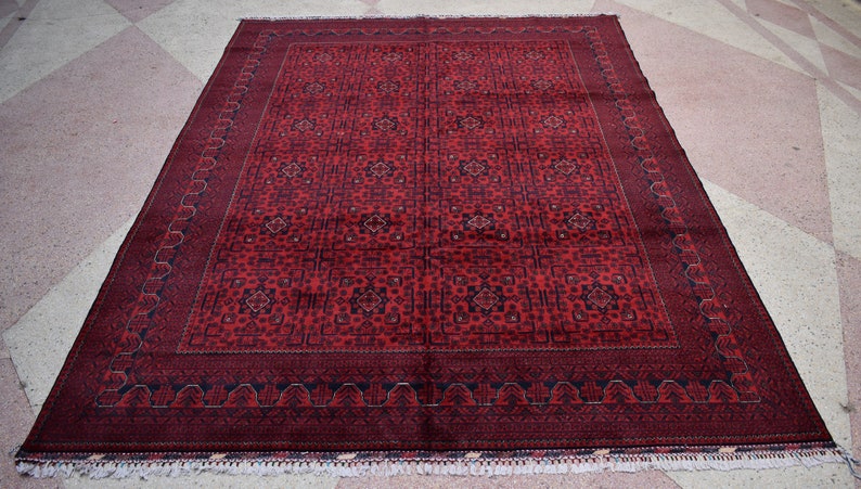 May include: A rectangular, hand-woven rug with a deep red field and intricate geometric patterns. The rug features a dark border and decorative fringe along the edges. The design includes repeating floral and geometric motifs.