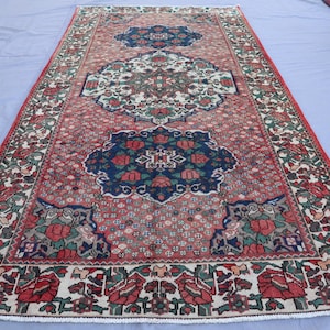 May include: A rectangular, vintage rug with a central medallion design. The rug features a red background with intricate floral patterns in white, blue, and green. The border is detailed with repeating floral motifs. The rug is likely made of wool.