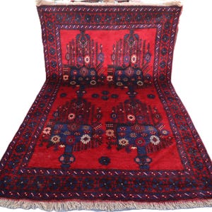 May include: A rectangular area rug featuring a vibrant red background with dark blue and white floral and geometric patterns. The rug has a wide border with repeating dark blue motifs and a fringe of light-colored tassels along the bottom edge.