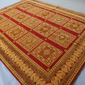 May include: A rectangular rug with a geometric pattern. The rug features a red field with repeating squares in shades of brown and gold. The border is a gold colour with repeating geometric shapes. The rug is made of a woven material.
