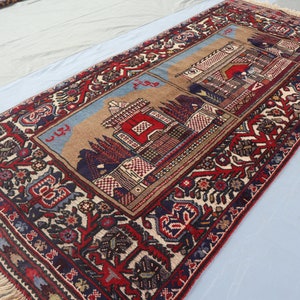 May include: A traditional Persian rug with a red, blue, and white floral pattern. The rug features a central design of a mosque with a red roof and a blue sky. The rug has a fringe border.