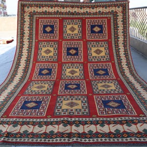 May include: A red, blue and cream coloured rug with a geometric pattern. The rug has a border with a repeating design of green leaves and cream flowers.
