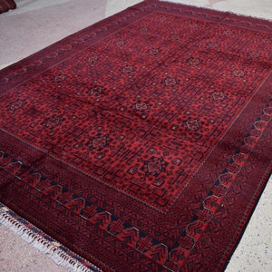 May include: A rectangular red rug with a detailed geometric pattern. The rug has a dark red border and a lighter red central field with repeating motifs. Fringes are visible along one edge. The rug's dimensions are not visible.