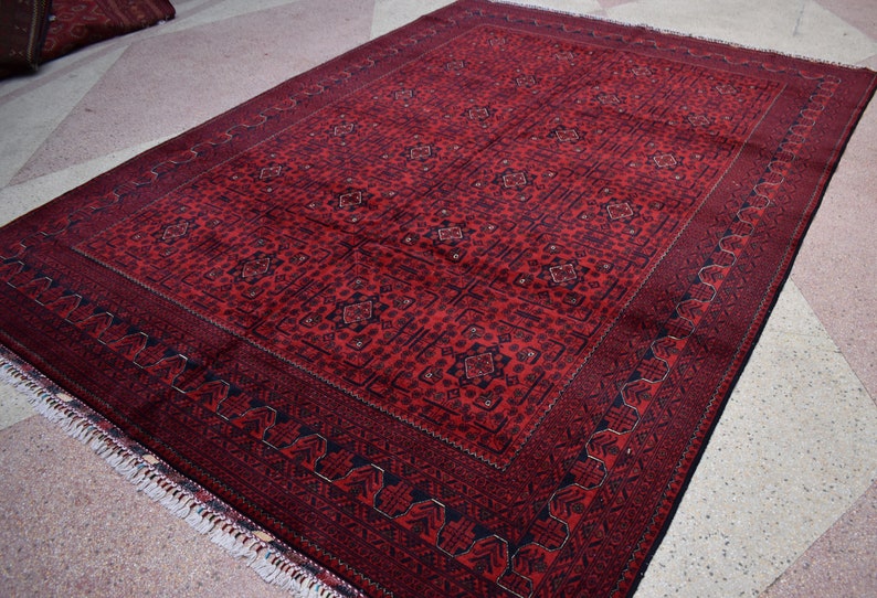 May include: A rectangular red rug with a detailed geometric pattern. The rug has a dark red border and a lighter red central field with repeating motifs. Fringes are visible along one edge. The rug's dimensions are not visible.