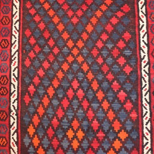 Pu&ograve; includere: Primo piano di un tappeto tessuto con un motivo geometrico. Il tappeto presenta un motivo centrale a rombi nei toni del rosso, arancione e blu, incorniciato da bordi con motivi ripetuti in rosso, bianco e blu. La trama del tappeto &egrave; visibile.