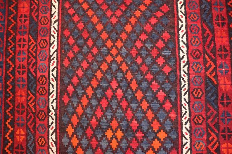 Pu&ograve; includere: Primo piano di un tappeto tessuto con un motivo geometrico. Il tappeto presenta un motivo centrale a rombi nei toni del rosso, arancione e blu, incorniciato da bordi con motivi ripetuti in rosso, bianco e blu. La trama del tappeto &egrave; visibile.