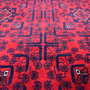 May include: A close-up of a red rug with a detailed geometric pattern. The rug features a central cross design and repeating floral and diamond shapes in navy blue and brown. The rug's texture is visible.