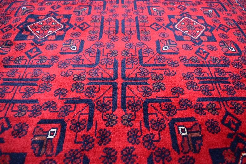 May include: A close-up of a red rug with a detailed geometric pattern. The rug features a central cross design and repeating floral and diamond shapes in navy blue and brown. The rug's texture is visible.
