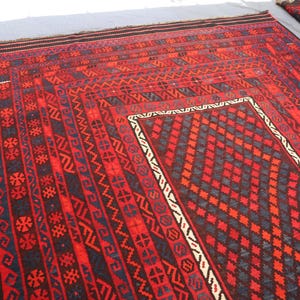 Pu&ograve; includere: Un grande tappeto rettangolare rosso con intricati motivi geometrici in nero, blu e bianco. Il tappeto presenta un disegno centrale a diamante e bordi con motivi ripetuti. Un tappeto pi&ugrave; piccolo e arrotolato &egrave; visibile sullo sfondo.