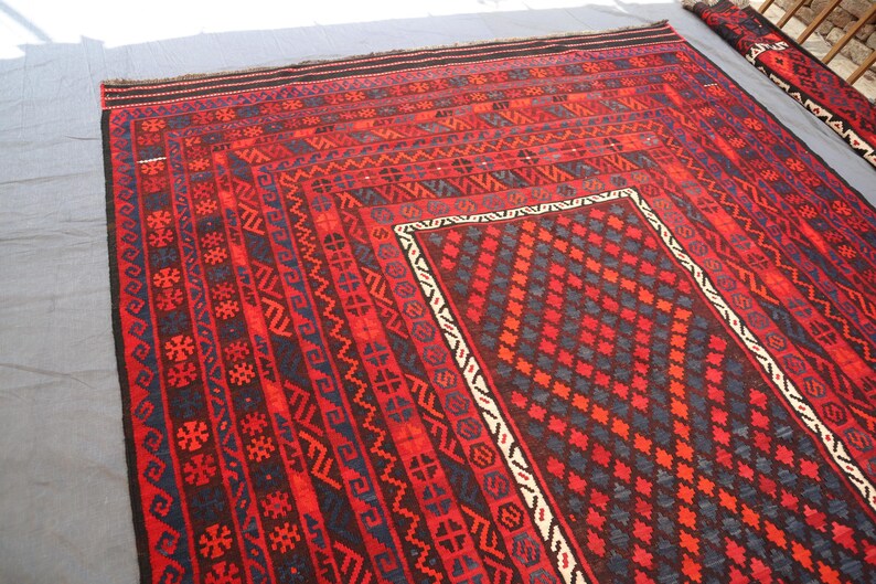 Pu&ograve; includere: Un grande tappeto rettangolare rosso con intricati motivi geometrici in nero, blu e bianco. Il tappeto presenta un disegno centrale a diamante e bordi con motivi ripetuti. Un tappeto pi&ugrave; piccolo e arrotolato &egrave; visibile sullo sfondo.