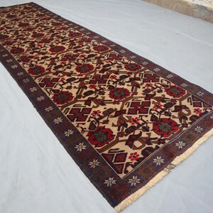May include: A long, rectangular runner rug featuring a beige field with a repeating pattern of red and brown floral and geometric designs. The border is dark brown with small white star-like shapes. The rug has a fringed edge.