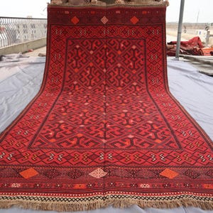 May include: A large, rectangular red rug with an intricate geometric pattern. The rug features a central design with repeating shapes and a border with diamond and geometric motifs. The rug has a fringe along the bottom edge.
