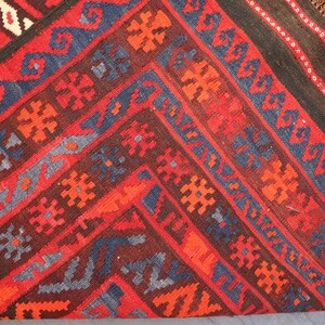 Pu&ograve; includere: Primo piano di un tappeto fantasia con un design geometrico. Il tappeto presenta una ricca tavolozza di colori di rosso, marrone, blu e arancione. I modelli e le trame intricati creano un tessuto visivamente accattivante.