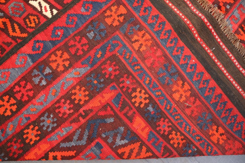 Pu&ograve; includere: Primo piano di un tappeto fantasia con un design geometrico. Il tappeto presenta una ricca tavolozza di colori di rosso, marrone, blu e arancione. I modelli e le trame intricati creano un tessuto visivamente accattivante.