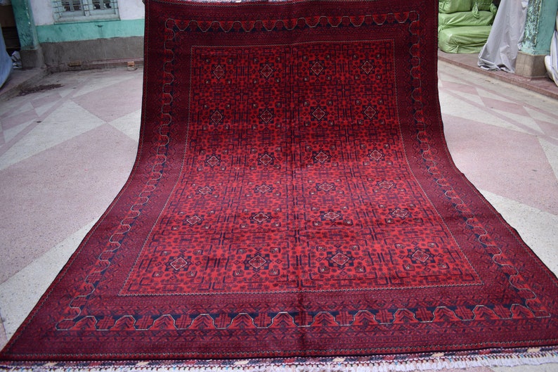 May include: A large, rectangular red rug with an intricate geometric pattern. The rug features a dark red border and a repeating design of squares and floral motifs. The rug is displayed on a light-colored surface.