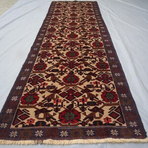 May include: A rectangular, beige runner rug with a repeating geometric pattern in red, brown, and dark blue. The rug features a central design of stylized floral and animal motifs, framed by a dark blue border with floral accents and fringe.