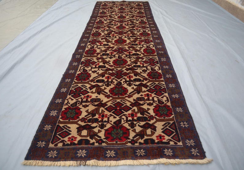 May include: A rectangular, beige runner rug with a repeating geometric pattern in red, brown, and dark blue. The rug features a central design of stylized floral and animal motifs, framed by a dark blue border with floral accents and fringe.