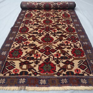 May include: Rolled-up beige runner rug with a detailed pattern. The design features red, brown, and green floral and geometric shapes. The border is dark blue with white star-like patterns and a cream fringe.