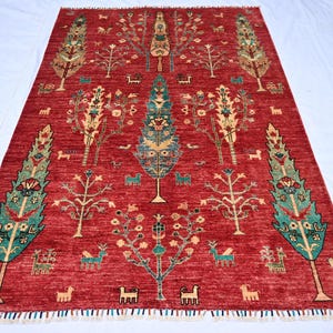 May include: A red, rectangular rug with a detailed tree and animal pattern. The rug features a central tree design with teal and gold accents, surrounded by smaller trees and animal figures. The rug has a fringe border.