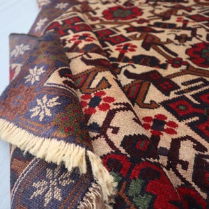 May include: A folded beige rug with a detailed pattern in red, brown, and blue. The design includes geometric shapes and floral motifs. The rug's edge features a blue border with white snowflake-like patterns and cream-coloured fringe.