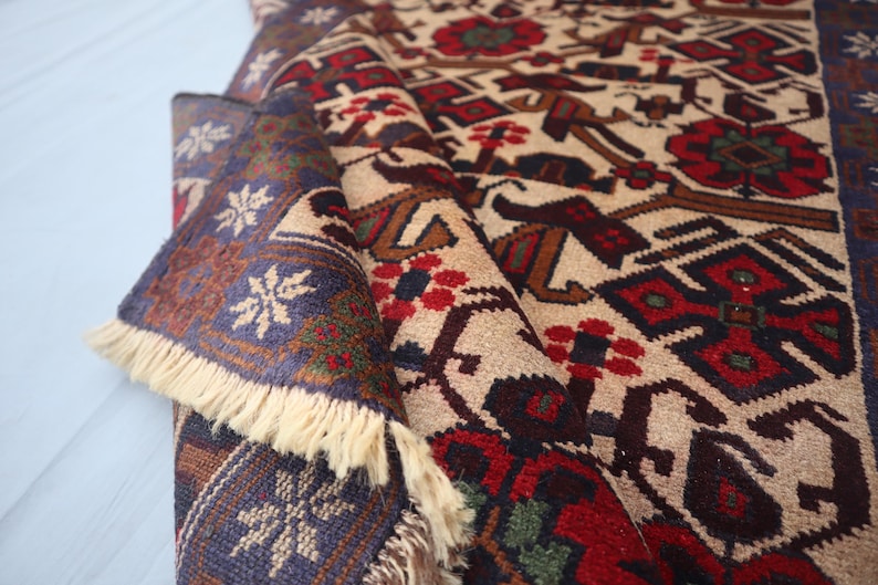May include: A folded beige rug with a detailed pattern in red, brown, and blue. The design includes geometric shapes and floral motifs. The rug's edge features a blue border with white snowflake-like patterns and cream-coloured fringe.