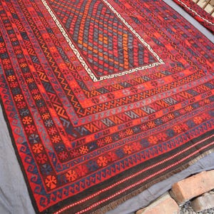 Pu&ograve; includere: Un tappeto rettangolare, tessuto a mano, con un motivo geometrico nelle tonalit&agrave; del rosso, nero, blu e arancione. Il tappeto presenta un disegno centrale a diamante e dettagli di bordo intricati. Il tappeto &egrave; esposto su una superficie grigia.