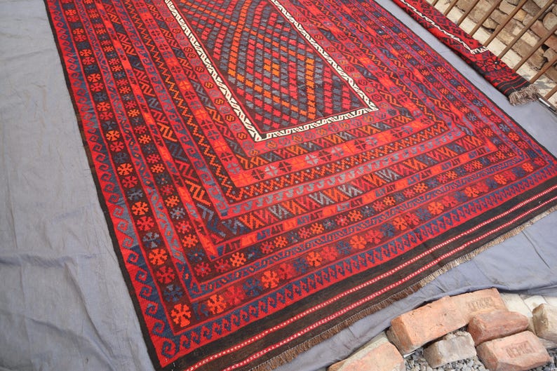 Pu&ograve; includere: Un tappeto rettangolare, tessuto a mano, con un motivo geometrico nelle tonalit&agrave; del rosso, nero, blu e arancione. Il tappeto presenta un disegno centrale a diamante e dettagli di bordo intricati. Il tappeto &egrave; esposto su una superficie grigia.