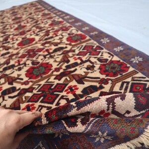 May include: A beige and red patterned rug with a dark blue border. The rug features floral and geometric designs in red, brown, and green. The rug is partially rolled up, revealing the underside.