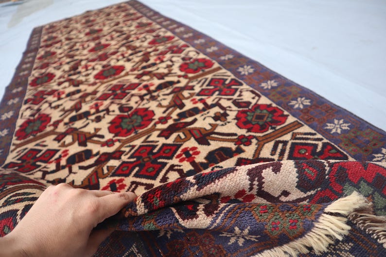 May include: A beige and red patterned rug with a dark blue border. The rug features floral and geometric designs in red, brown, and green. The rug is partially rolled up, revealing the underside.
