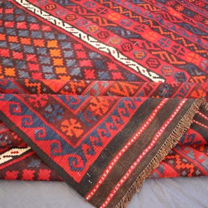 Pu&ograve; includere: Un tappeto piegato con un motivo geometrico rosso, nero e blu. Il tappeto presenta disegni a rombi, zigzag e floreali. Ha un bordo marrone e frange. Il design &egrave; intricato e dettagliato.