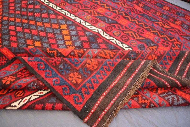 Pu&ograve; includere: Un tappeto piegato con un motivo geometrico rosso, nero e blu. Il tappeto presenta disegni a rombi, zigzag e floreali. Ha un bordo marrone e frange. Il design &egrave; intricato e dettagliato.