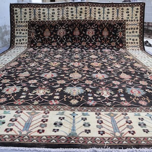 May include: A large, rectangular rug with an intricate floral pattern in shades of black, beige, red, and blue. The rug features a central dark field with a repeating floral design and a beige border with stylized floral and geometric motifs.