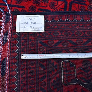 May include: A close-up of a red and black handmade rug with a geometric pattern. The rug has a fringe edge and a tag with handwritten details. A measuring tape shows the rug's width. The rug's size is 11.9 x 8.3 inches.