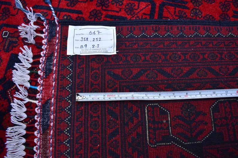 May include: A close-up of a red and black handmade rug with a geometric pattern. The rug has a fringe edge and a tag with handwritten details. A measuring tape shows the rug's width. The rug's size is 11.9 x 8.3 inches.
