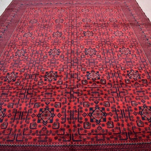May include: A large, rectangular red rug with an intricate geometric pattern. The rug features a repeating design of squares, diamonds, and floral motifs in dark blue and red. The border is a darker shade of red with a repeating pattern.
