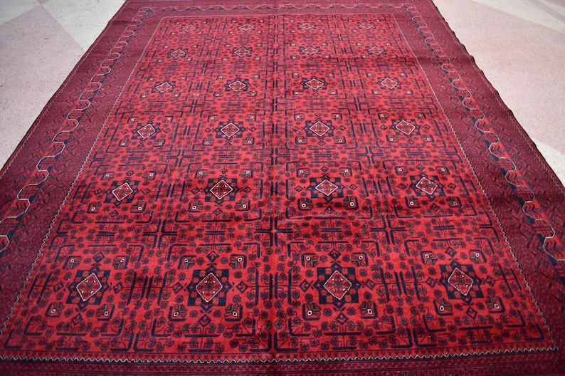 May include: A large, rectangular red rug with an intricate geometric pattern. The rug features a repeating design of squares, diamonds, and floral motifs in dark blue and red. The border is a darker shade of red with a repeating pattern.