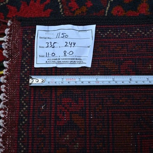 Puede incluir: Primer plano de una alfombra roja y negra con flecos blancos. Una etiqueta blanca con escritura negra est&aacute; unida a la alfombra. La etiqueta dice "Serial No. 1150, Size: 335 x 244, Size: 11.0 x 8.0,  D OF HANDMADE RUGS & KILIMS 130% HAND SPUN WOOL". Una regla de metal se coloca sobre la alfombra.