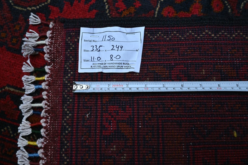 Puede incluir: Primer plano de una alfombra roja y negra con flecos blancos. Una etiqueta blanca con escritura negra est&aacute; unida a la alfombra. La etiqueta dice "Serial No. 1150, Size: 335 x 244, Size: 11.0 x 8.0,  D OF HANDMADE RUGS & KILIMS 130% HAND SPUN WOOL". Una regla de metal se coloca sobre la alfombra.