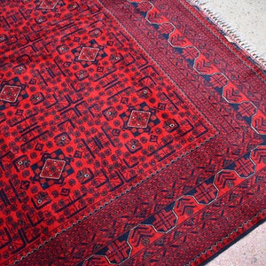 May include: A red and navy blue patterned rug with a geometric design. The rug features a central field with repeating diamond and floral motifs, framed by a detailed border. The rug's edges have a fringe.