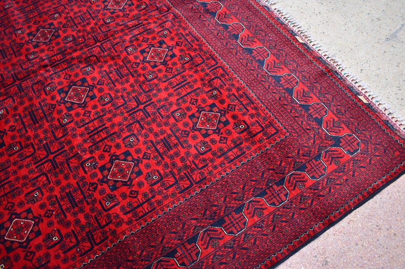 May include: A red and navy blue patterned rug with a geometric design. The rug features a central field with repeating diamond and floral motifs, framed by a detailed border. The rug's edges have a fringe.