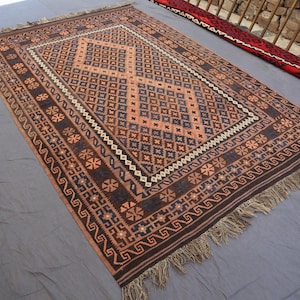 May include: A rectangular rug featuring a geometric design in brown, tan, and navy blue. The central pattern is diamond-shaped, with floral and geometric borders. Fringes are present along the edges of the rug.