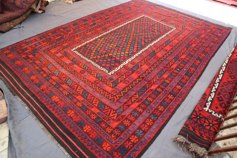 Pu&ograve; includere: Un grande tappeto rettangolare rosso con un motivo geometrico centrale nelle tonalit&agrave; dell'arancione, del blu e del nero. Il tappeto presenta intricati motivi a bordo e un corridore abbinato. Il tappeto &egrave; esposto su una superficie grigia.
