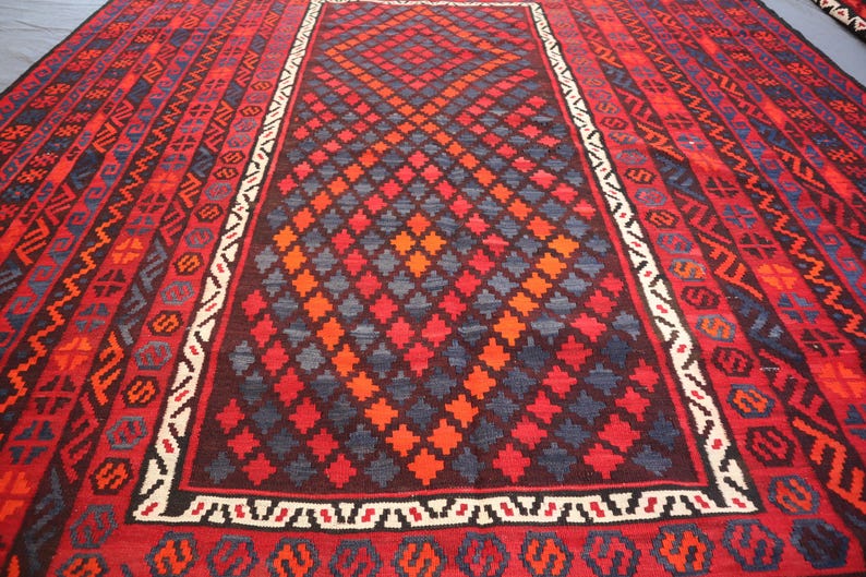 Pu&ograve; includere: Un grande tappeto rettangolare con un motivo a diamante centrale in rosso, arancione e blu. Il bordo presenta disegni geometrici in rosso, nero e bianco. La combinazione di colori del tappeto &egrave; prevalentemente rossa.