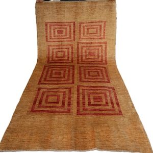 May include: A large, rectangular rug with a tan background and a repeating pattern of red squares. The rug is made of a woven material and has a soft, textured appearance. The design features six large squares in a grid formation.